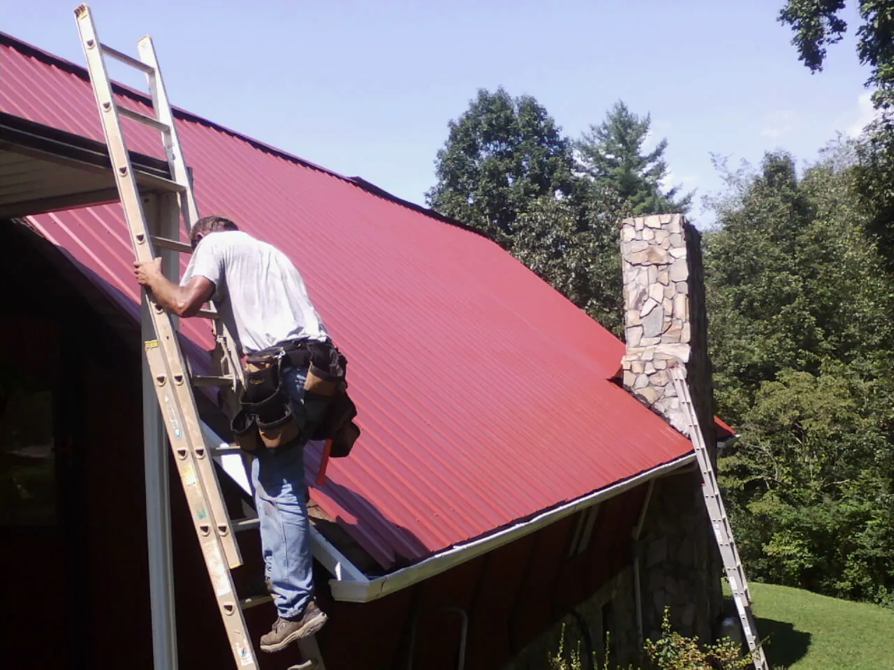 Roofer on a ladder beside a red metal roof and stone chimney — Soffit & Fascia Repair by Roofing Brothers in Dubuque